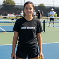 Woman on a tennis court wearing a black t-shirt with 'JUST DROP IT.' text, holding a tennis ball and racket.

